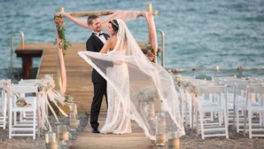 Uma cerimônia de casamento na praia, com um píer de madeira, cadeiras brancas e velas.