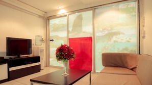 A living room with a television, a coffee table with a vase of flowers, a sofa, and a large window with a view of a mountainous landscape. - Hello Backpacker Samui Hostel (Koh Samui)