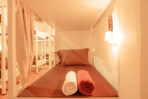 A single bed with a brown mattress, two rolled towels (one white, one red), and a white wall with a light fixture.