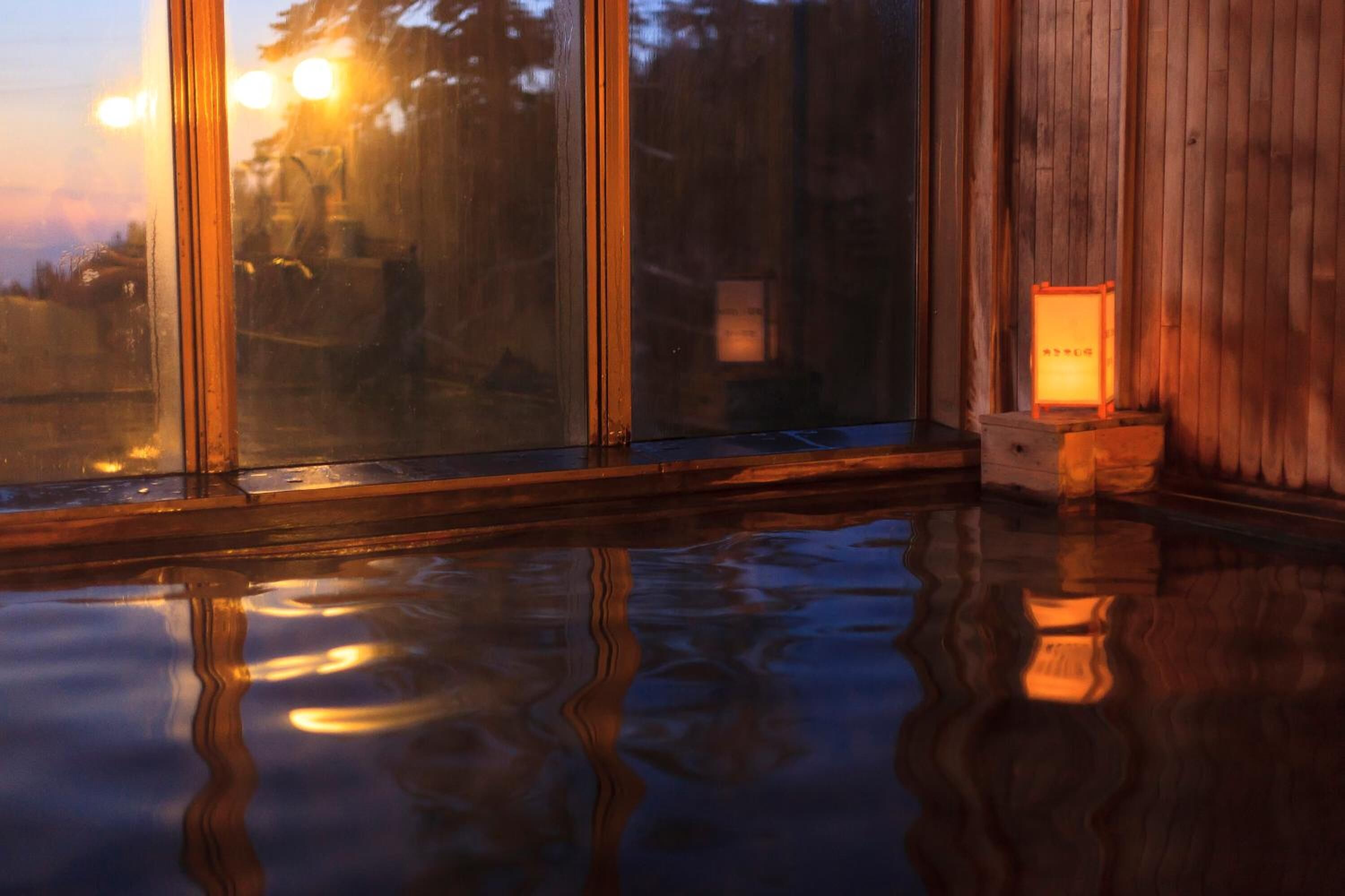 A room with a large window reflecting a sunset, a wooden floor, and a small table with a lamp.