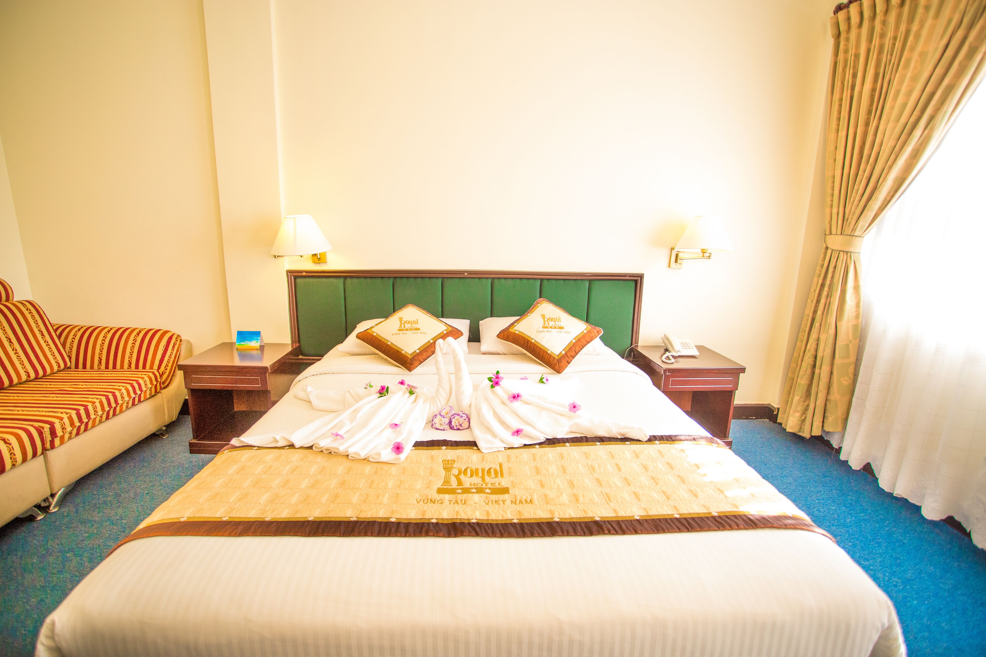 A hotel room with a neatly made bed, a sofa, a bedside table with a lamp, and a window with curtains.