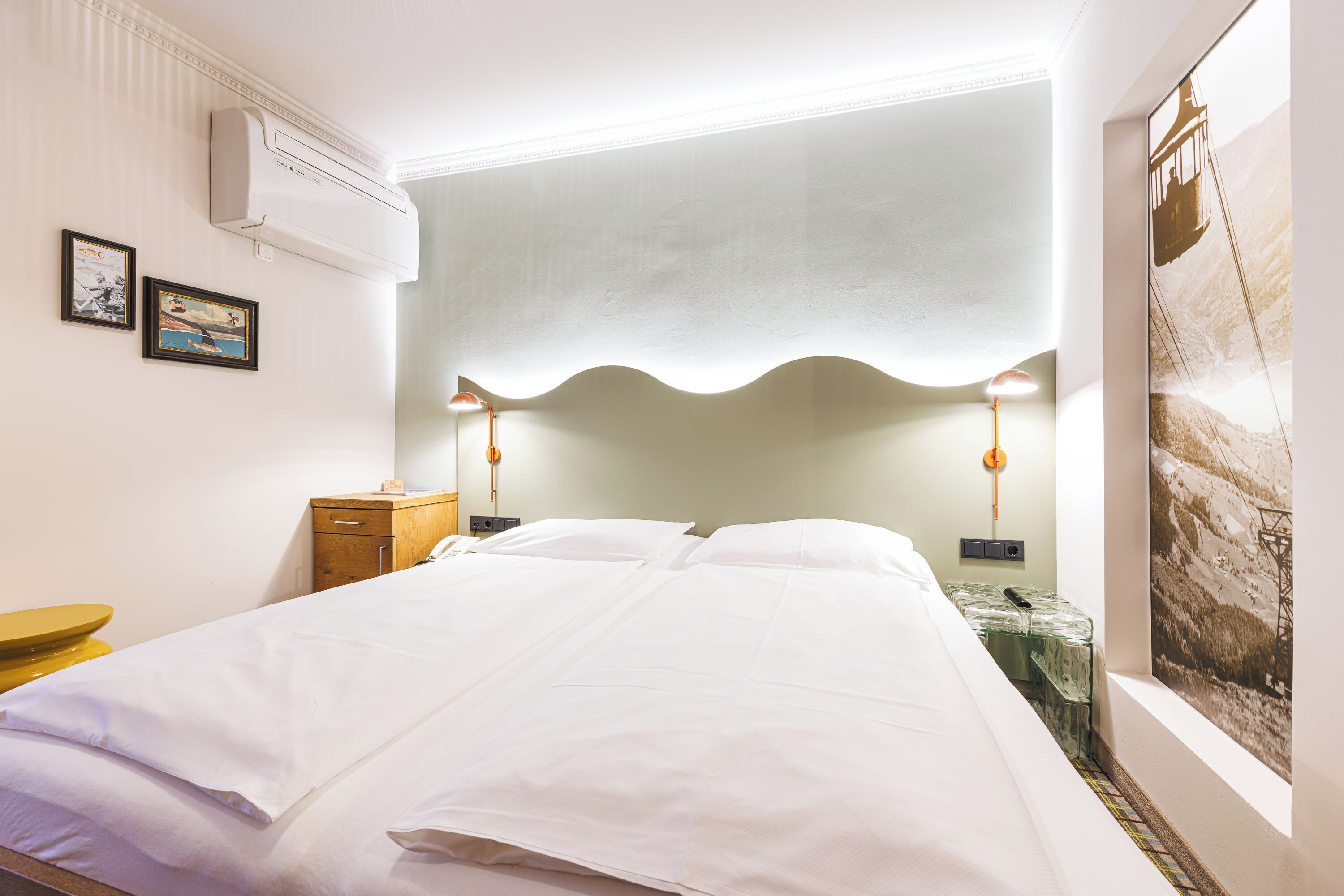 A neatly made bed with white linens, a bedside table with a lamp, and a wall-mounted air conditioner.