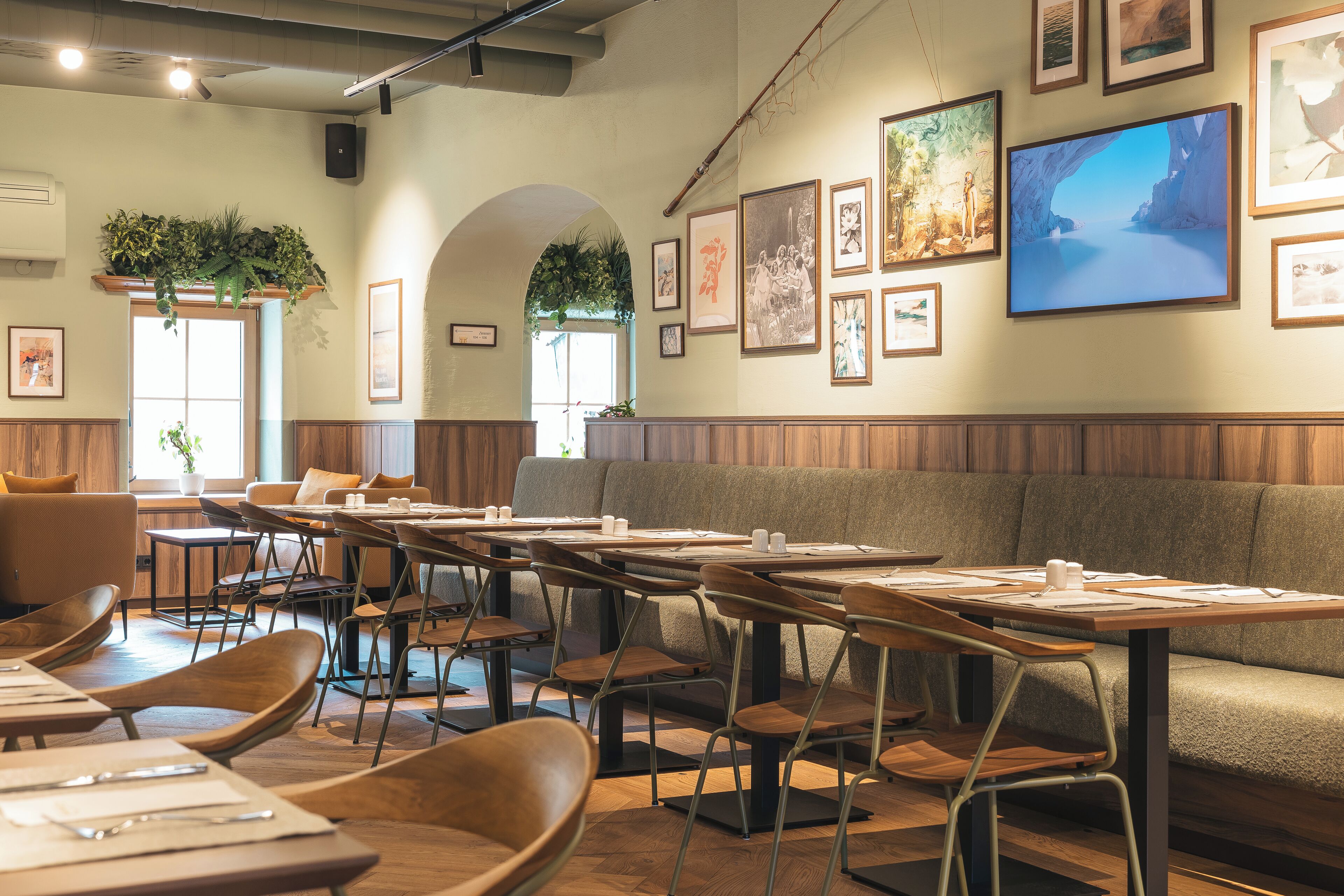 A restaurant with booths, tables, and chairs. There are framed pictures on the walls and a large window with plants.