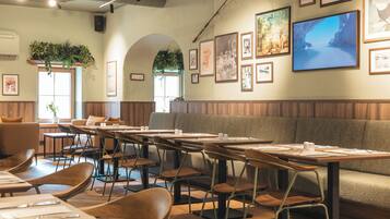 Un restaurant avec des banquettes, des tables et des chaises. Les murs sont ornés de tableaux encadrés et il y a une grande fenêtre avec des plantes.