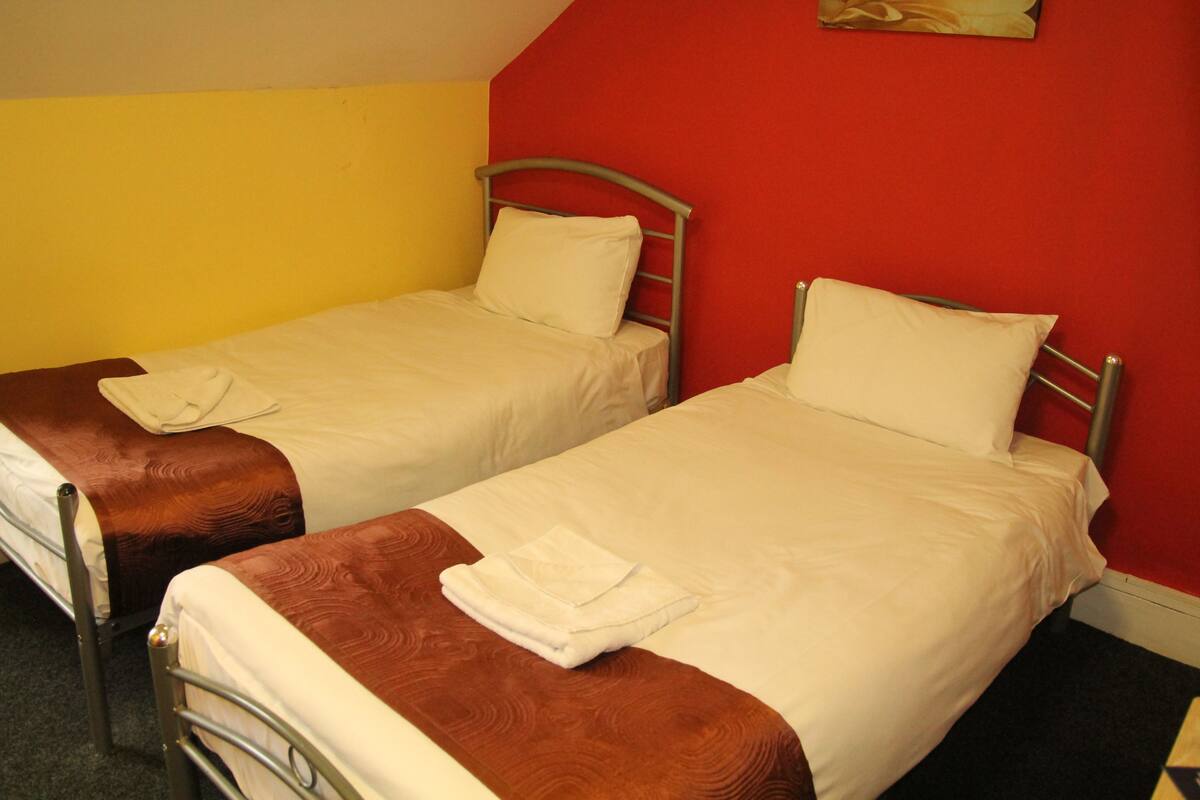 Deux lits simples avec des structures en métal, du linge de lit blanc et des têtes de lit marron, dans une pièce aux murs jaunes et rouges.