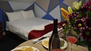 Une chambre d’hôtel avec un lit, une table à manger dressée avec des assiettes de nourriture, une bouteille de vin et un vase rempli de fleurs.