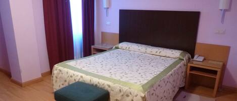 Habitación de hotel con cama, mesita de noche, reposapiés verde y ventana con cortinas.