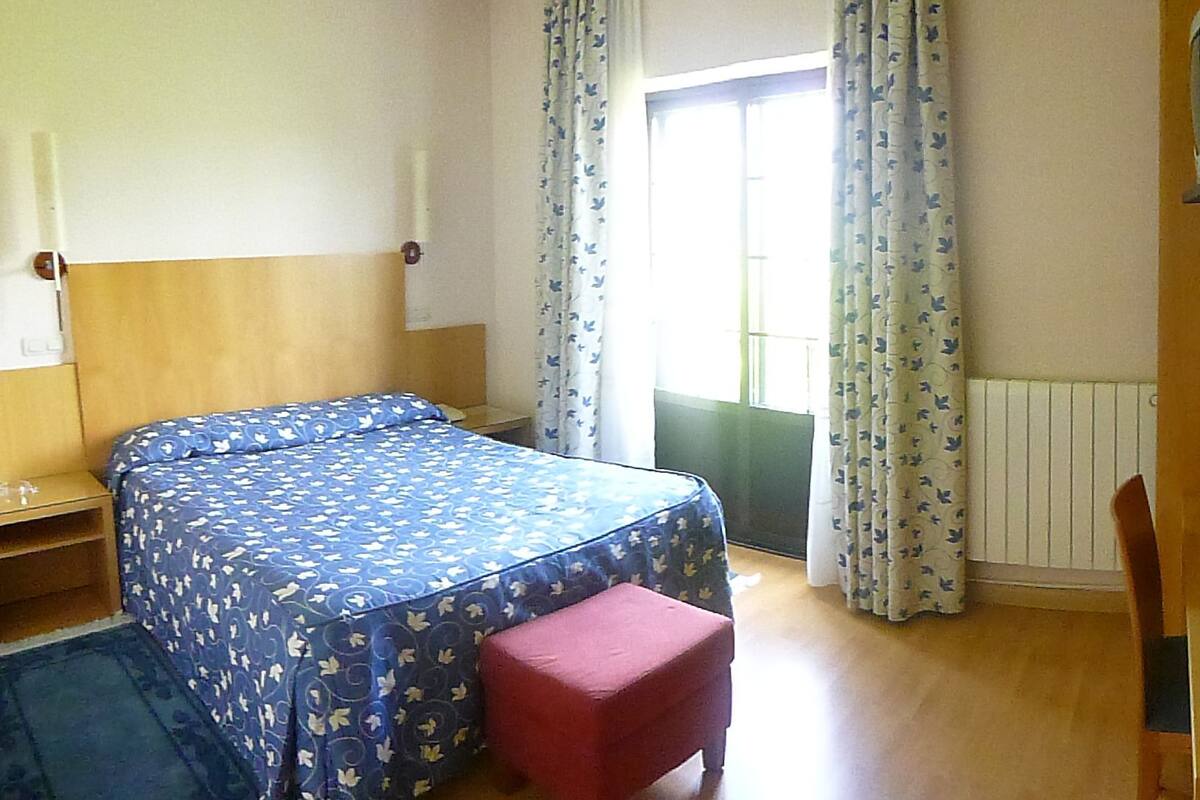 A bedroom with two beds, a red bench, a window with curtains, and a radiator.
