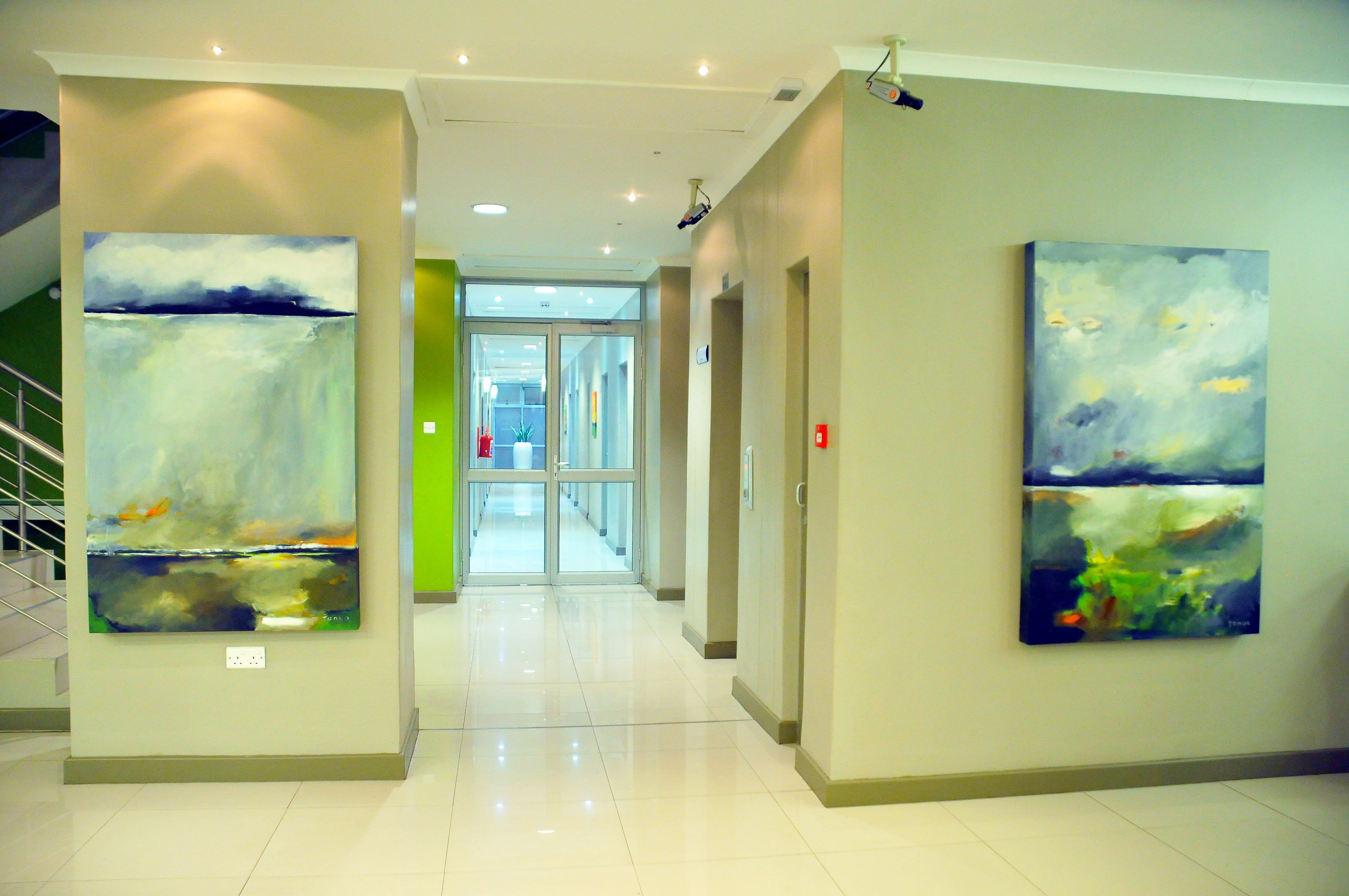 A hallway with abstract paintings on the walls, a staircase to the left, and a door at the end.