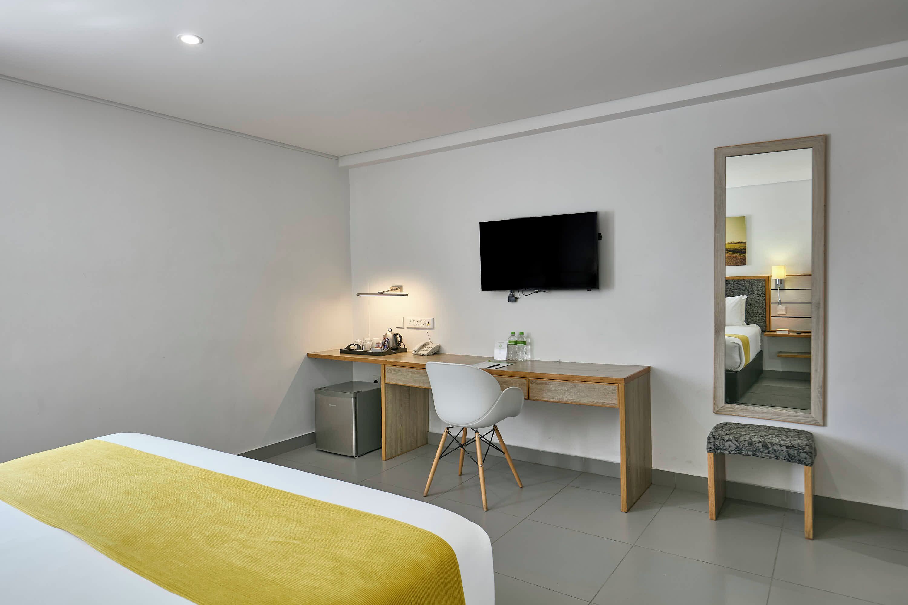 A modern hotel room with a bed, a desk with a chair, a television, and a large mirror.