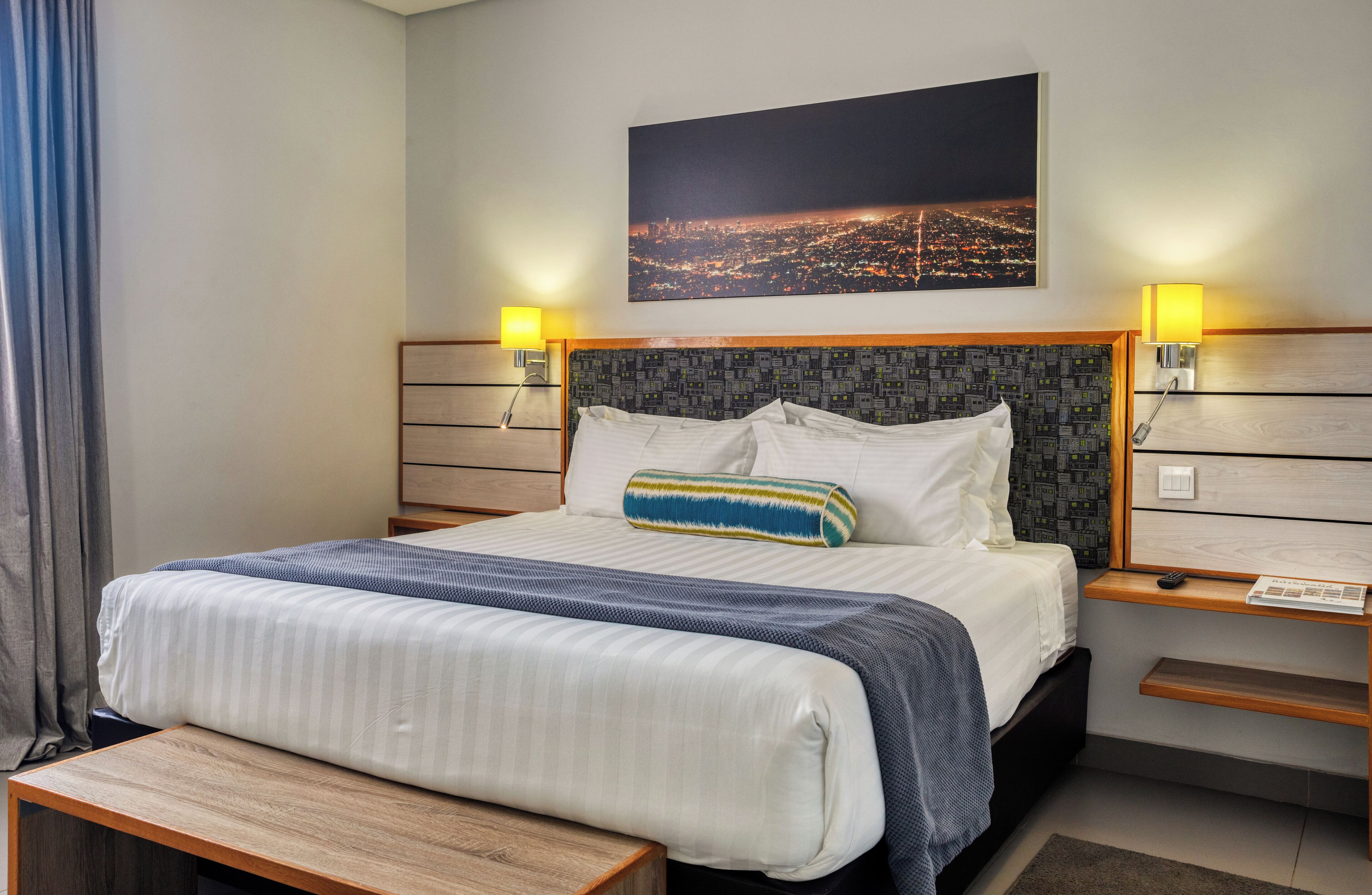 Hotellihuone, jossa on suuri sänky, kaksi yöpöytää, yölamppu, penkki, näköala kaupunkiin ja kaupungin valojen kuva seinällä.