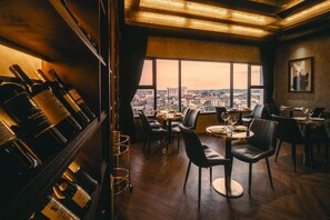 Un restaurante con vistas a un paisaje urbano, suelos de madera y una estantería con botellas de vino.