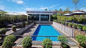 Un área de piscina en la azotea con sillones de descanso, plantas en macetas y un edificio moderno al fondo.