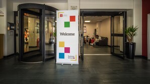 Una entrada de edificio moderna con puerta de vidrio, cartel de bienvenida y planta en maceta.