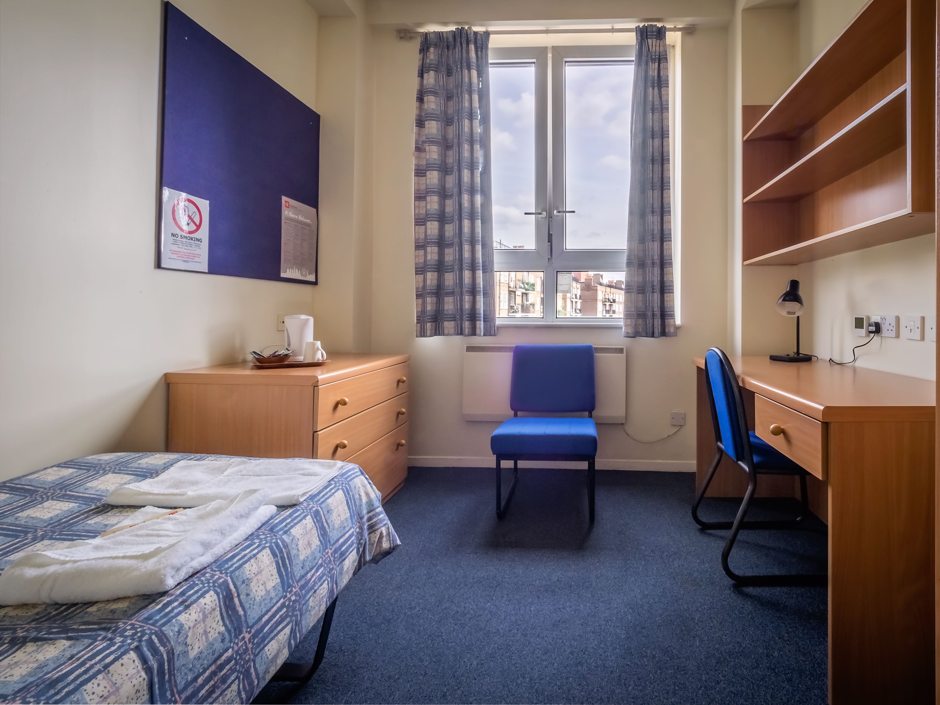 A small, tidy room with a bed, a desk, a chair, and a window with curtains.