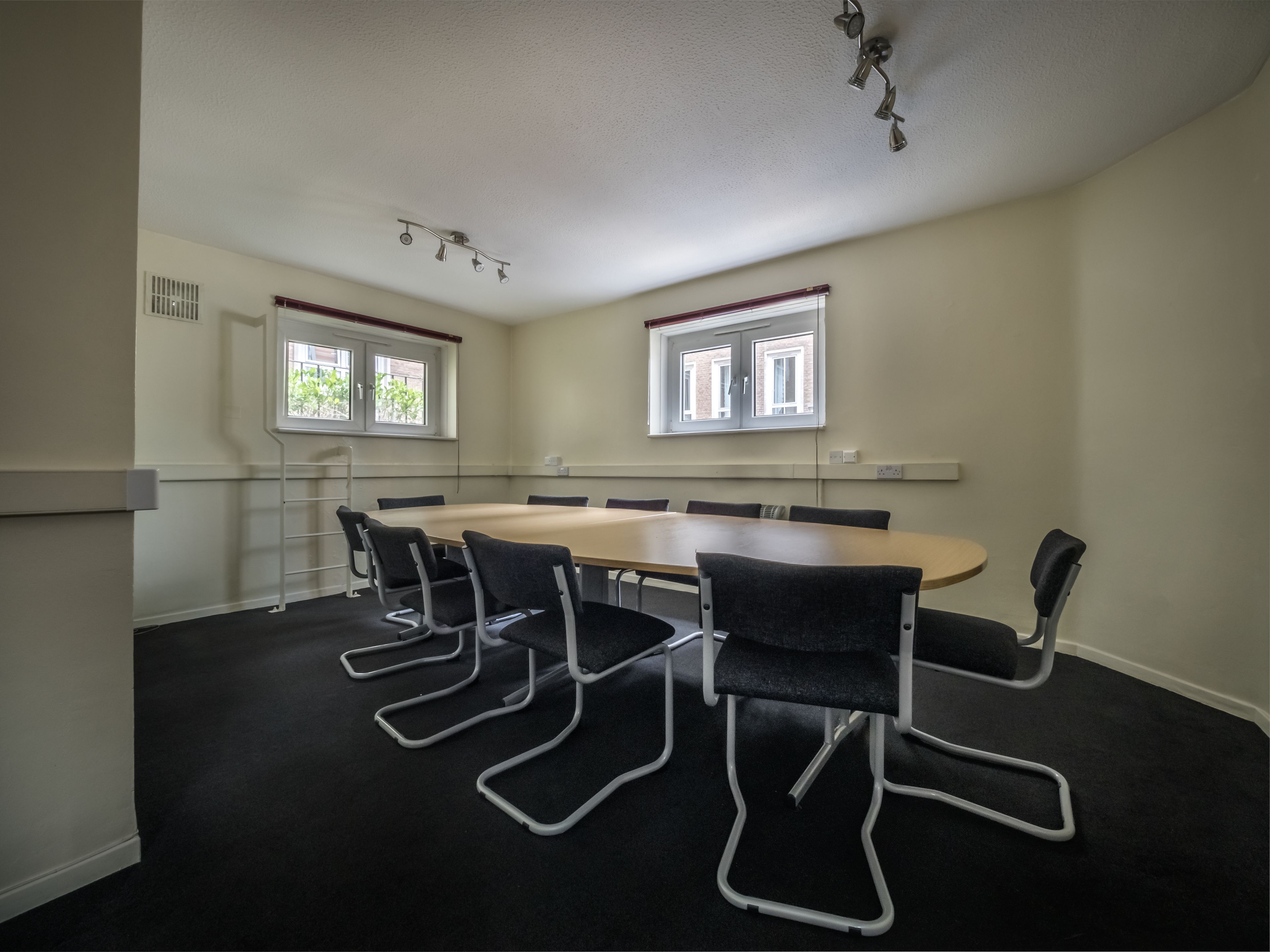Ein Konferenzraum mit einem langen Tisch, sechs Stühlen, zwei Fenstern und einer Deckenleuchte.