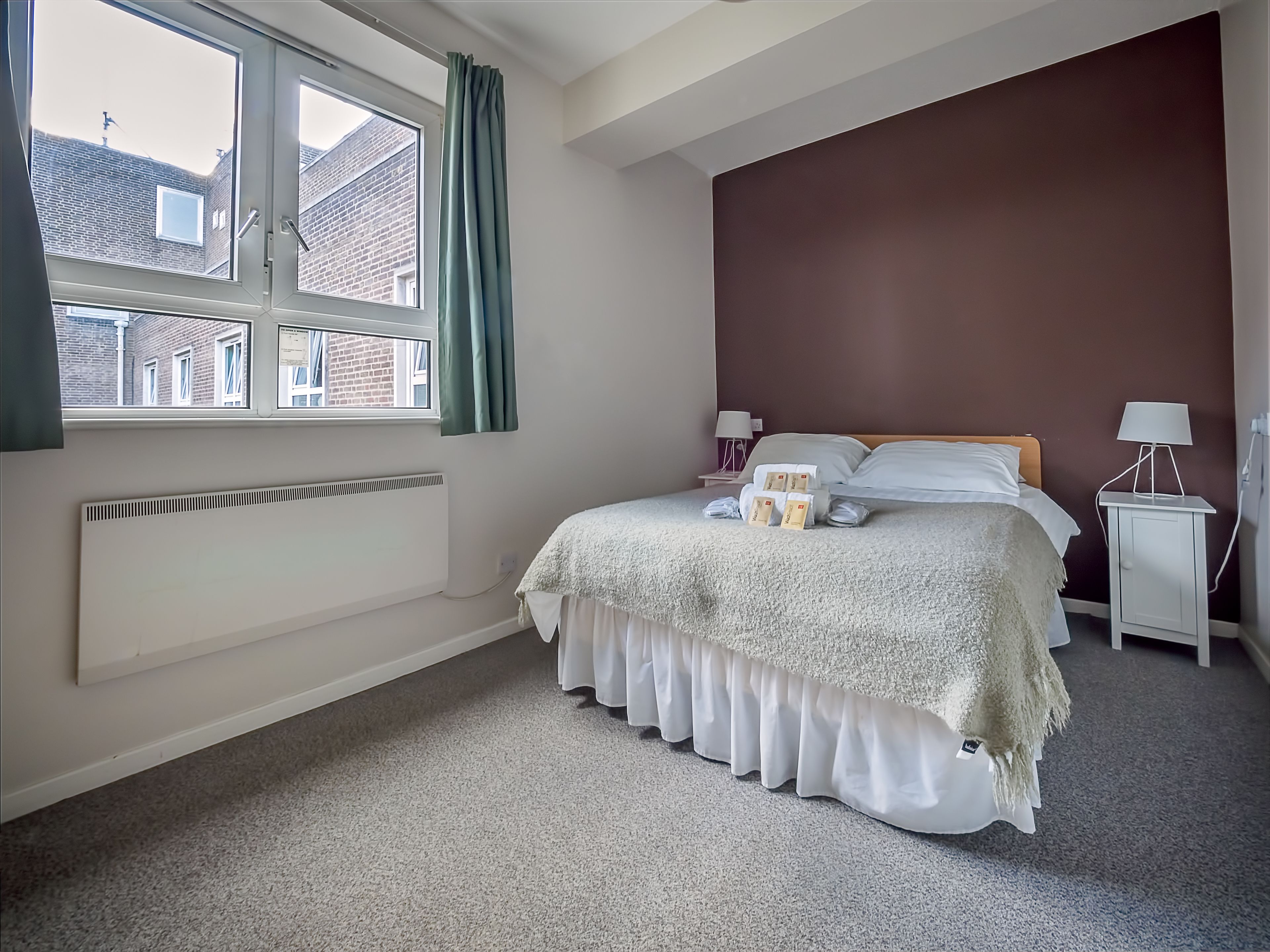 A bedroom with a bed, bedside tables, a window with curtains, a radiator, and a view of buildings outside.