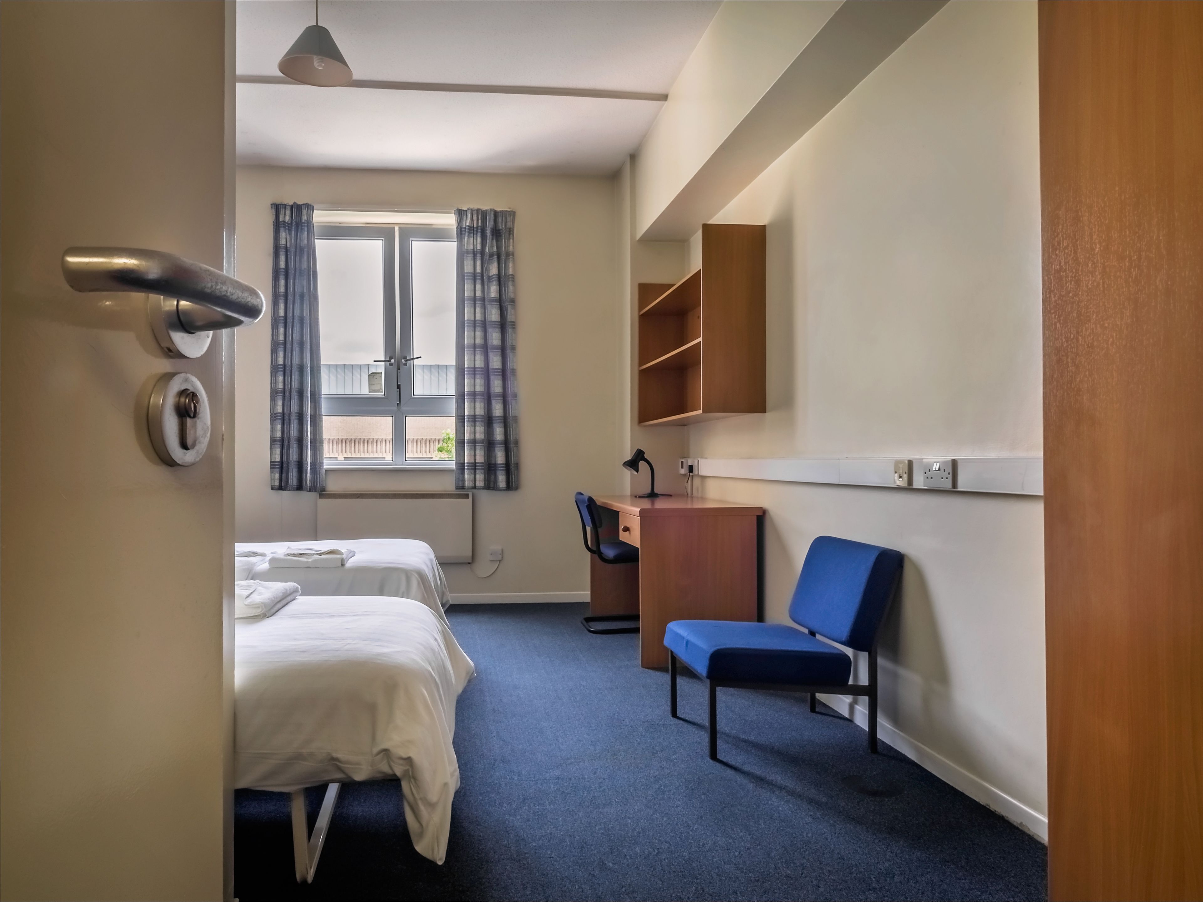 Habitación de hotel con cama, escritorio con silla, ventana con cortinas y estante en la pared.