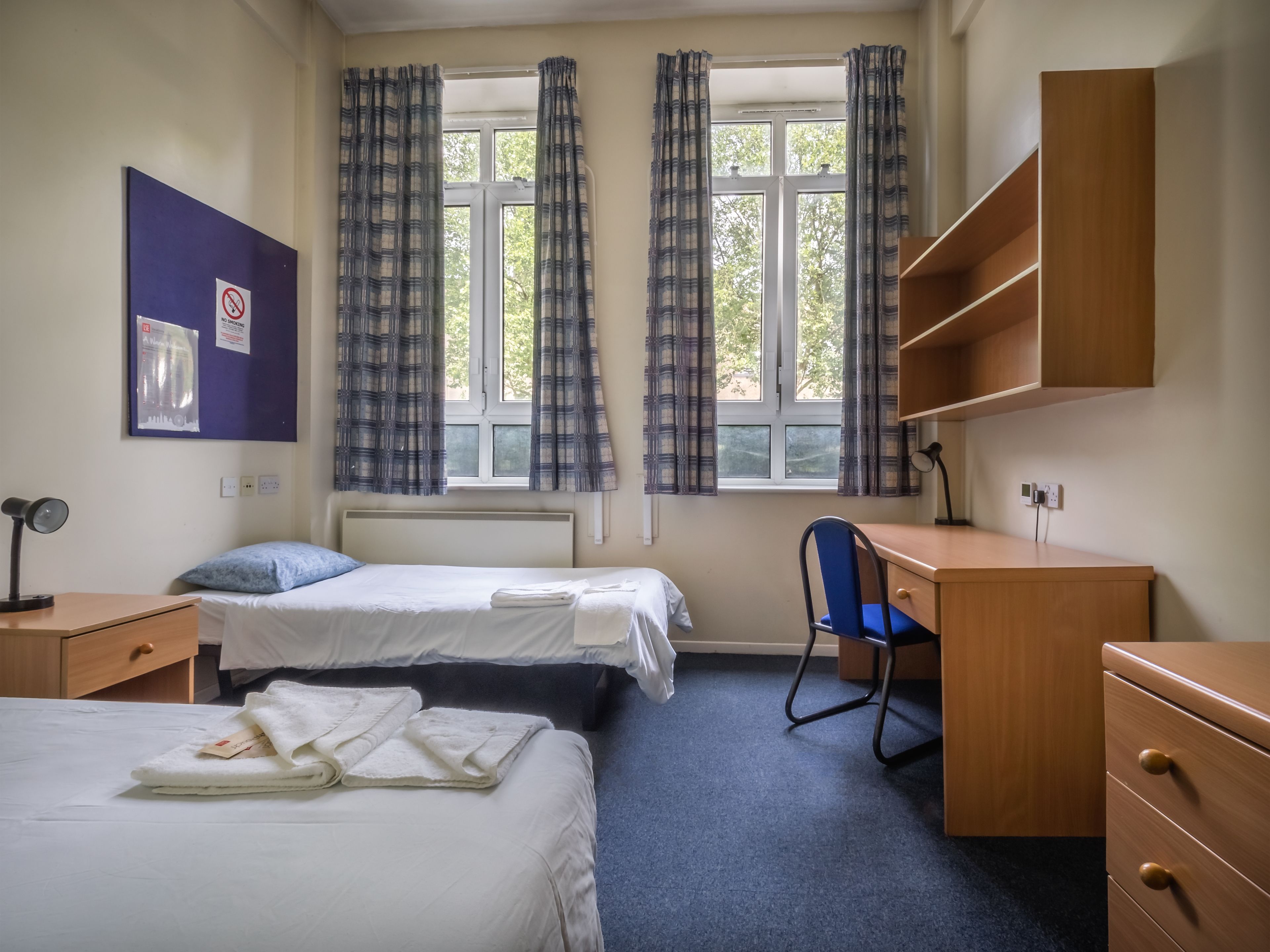 A dormitory room with two beds, a desk, a chair, and a shelf.