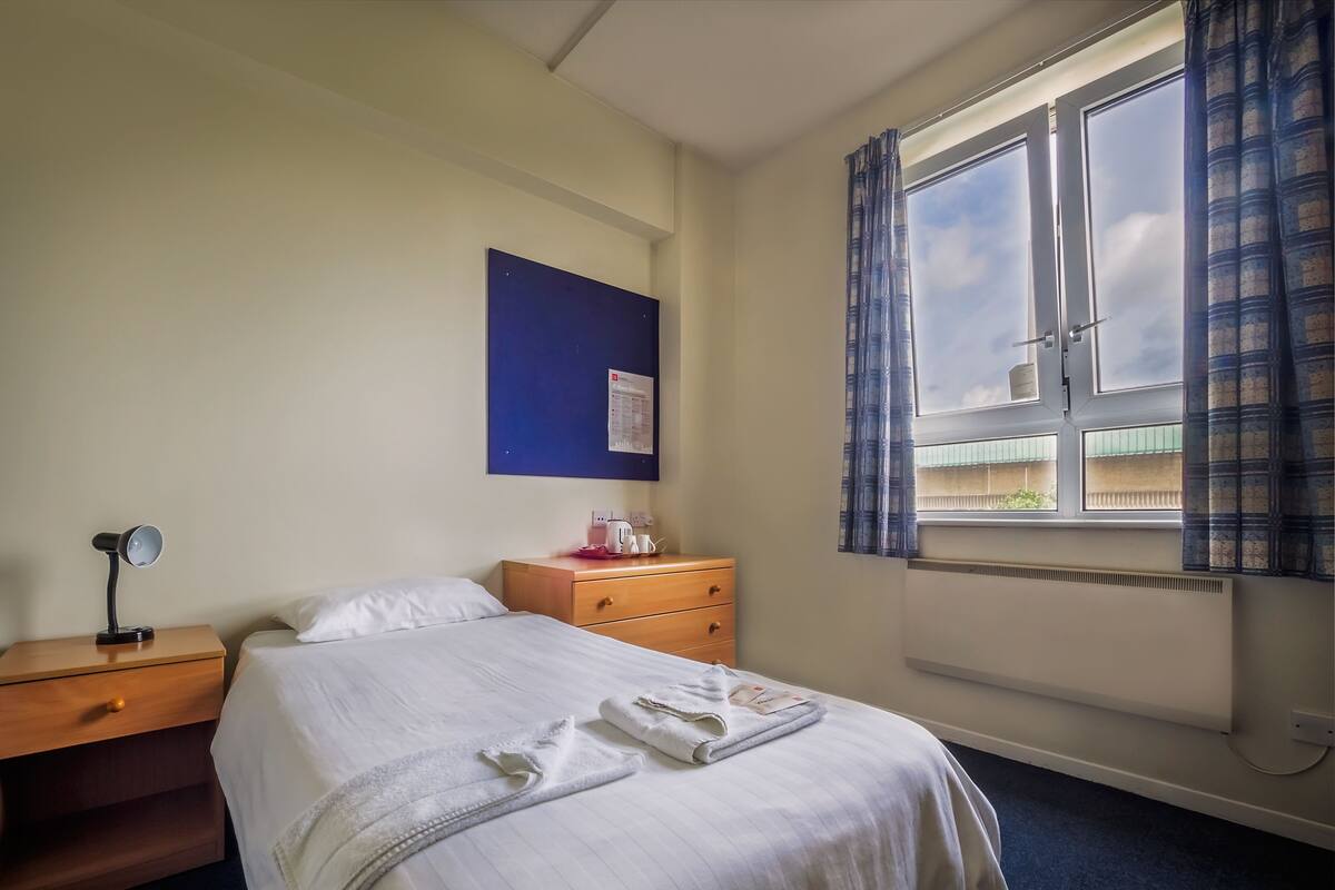 Hotellihuone, jossa on sänky, yöpöytä, työpöytä, ikkuna verhoilla ja seinälle kiinnitetty ilmoitustaulu.