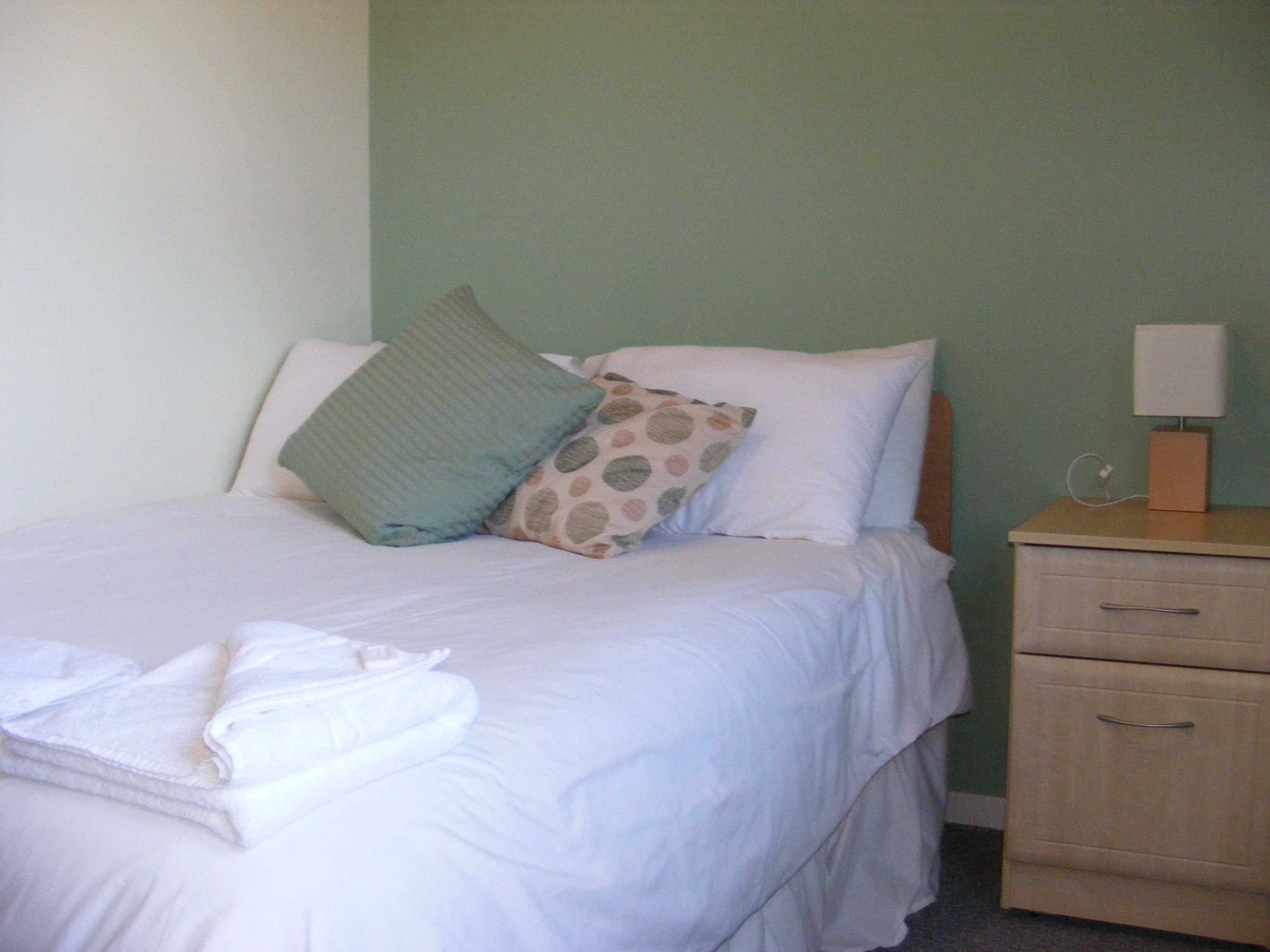 A neatly made bed with a pillow, a bedside table with a lamp, and a drawer.