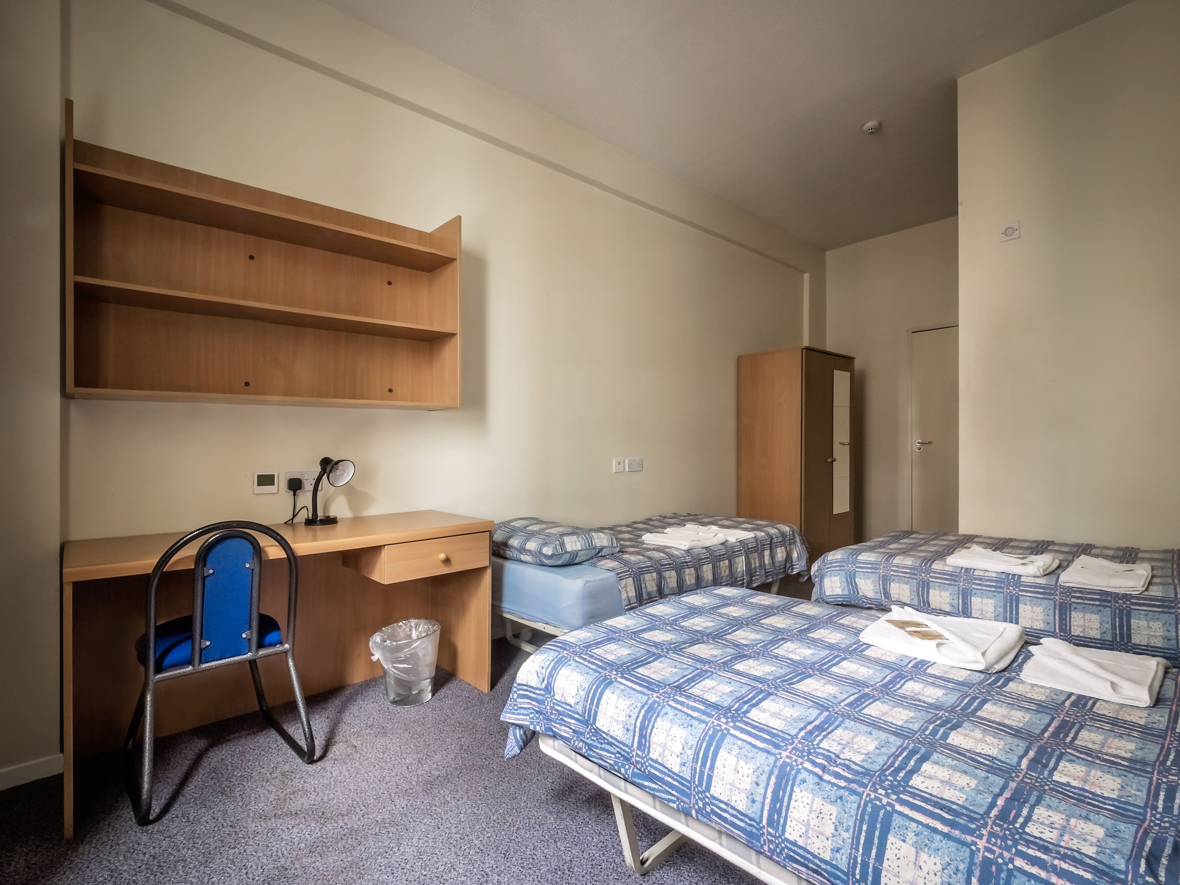 Una habitación con escritorio, dos camas con ropa de cama estampada en azul, un armario y una silla con cojín azul.