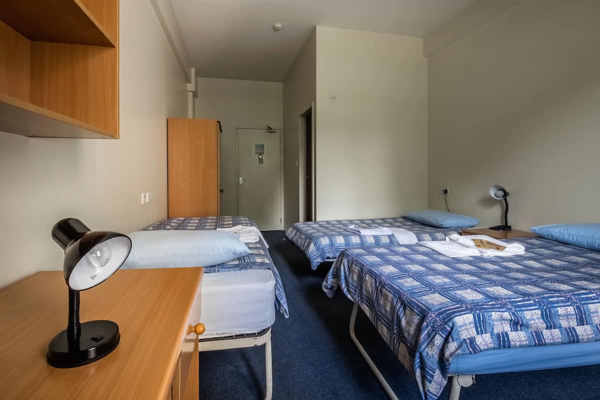 Hotellihuone, jossa on kaksi sänkyä, työpöytä lampun kanssa, vaatekaappi ja ovi, joka johtaa toiseen huoneeseen.