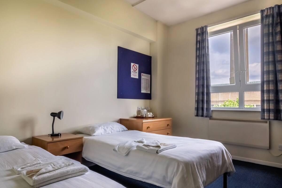 Hotellihuone, jossa on kaksi sänkyä, yöpöytä, työpöytä, ikkuna verhoilla ja seinälle kiinnitetty kyltti.