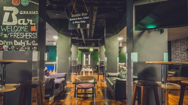 Zona de bar con taburetes, un sofá y un letrero que indica un bar de botellas que sirve cerveza, sidra y vino embotellados.