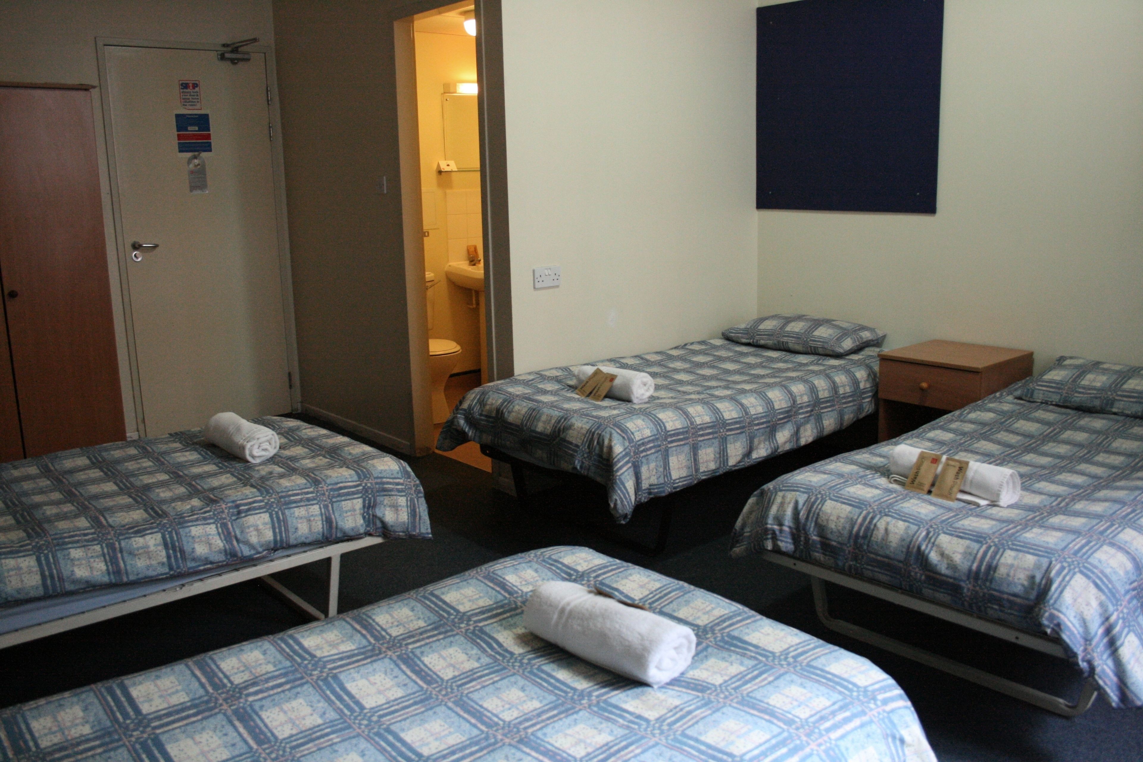 A hotel room with three single beds, each with a blue and white checkered bedspread and a rolled towel on the bed. There is a wooden door on the left, a bathroom visible through an open door, and a dark blue wall on the right.