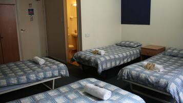 A hotel room with three single beds, each with a blue and white checkered bedspread and a rolled towel on the bed. There is a wooden door on the left, a bathroom visible through an open door, and a dark blue wall on the right.