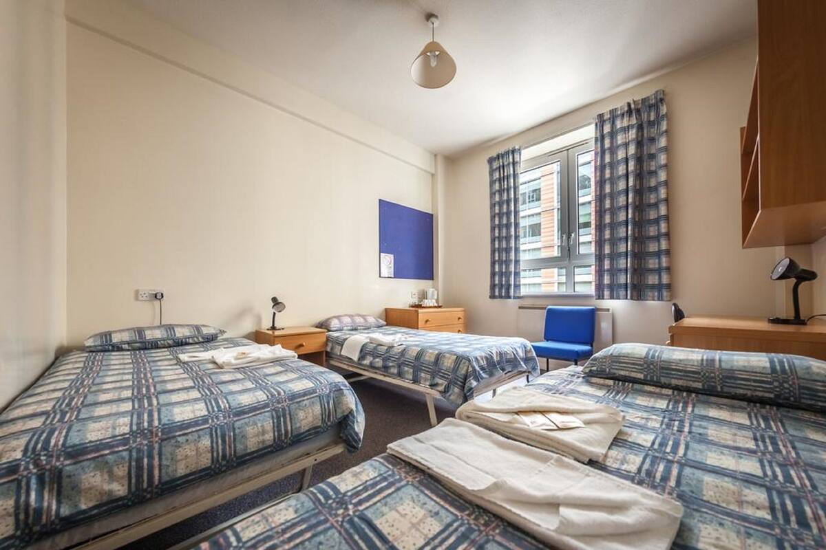 Hotellihuone, jossa on kolme sänkyä, työpöytä, tuoli, ikkuna, jossa on verhot, ja sininen seinäkoriste.