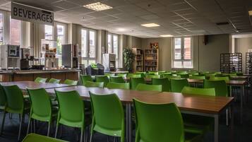 Une cafétéria avec des chaises et des tables vertes, un espace dédié aux boissons et une bibliothèque.