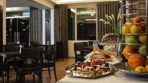 Un bufé de desayuno con frutas, pasteles y bebidas en un comedor de hotel con decoración moderna y ventanas amplias.