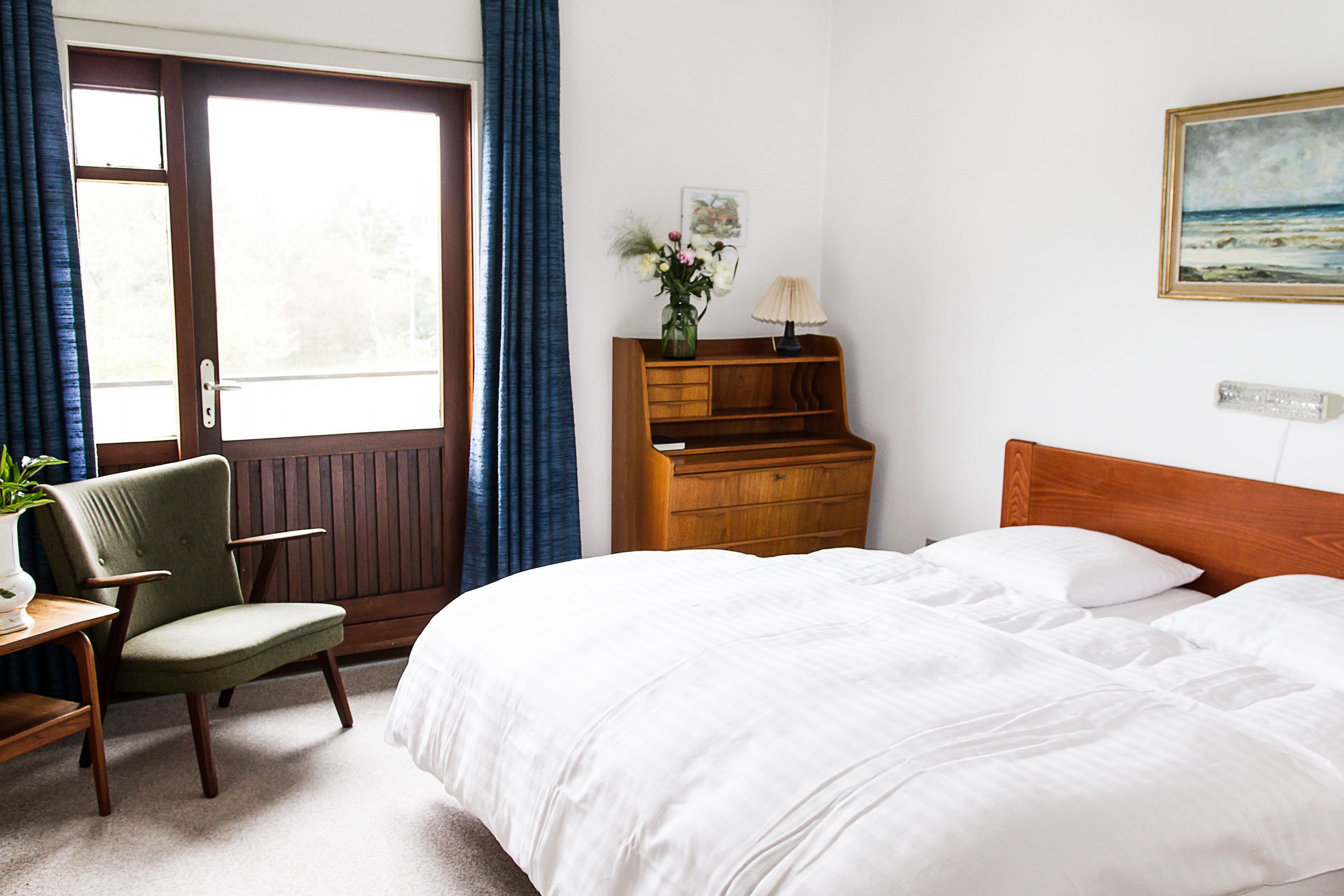 A hotel room with a bed, a chair, a desk, a dresser, a window with curtains, and a painting on the wall.