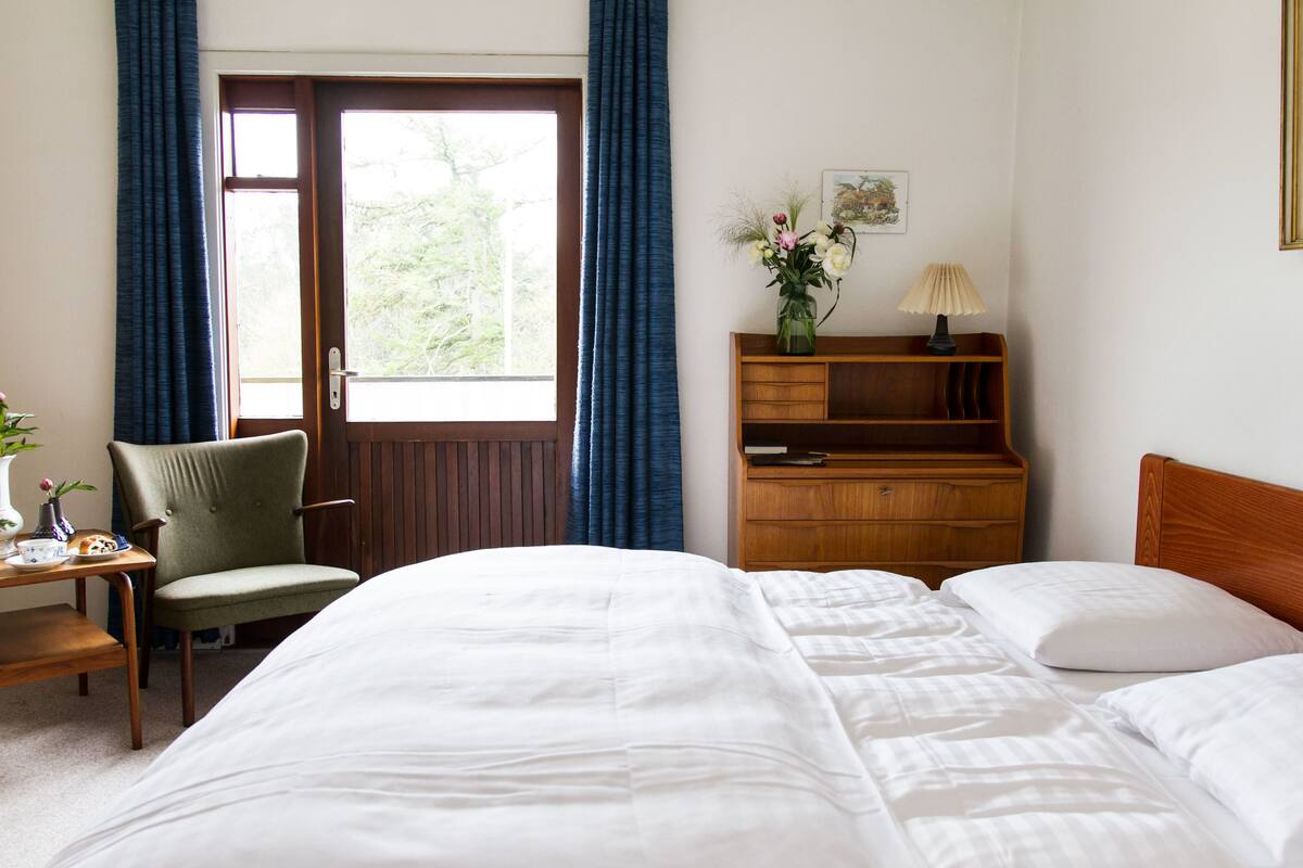 A hotel room with a large bed, a wooden nightstand, a chair, a desk with a lamp, and a window with curtains.