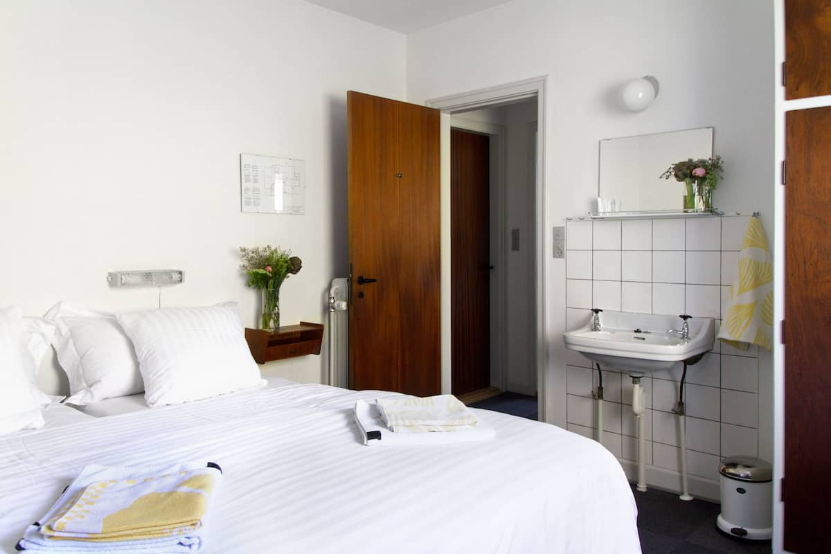 A hotel room with a neatly made bed, a wooden door, a white sink, and a mirror above it.