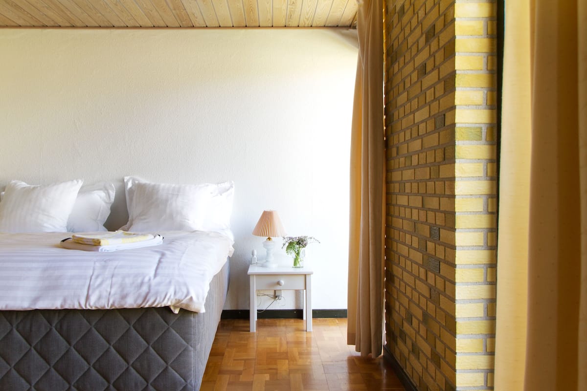A bedroom with a bed, a bedside table with a lamp and vase, and a brick wall.