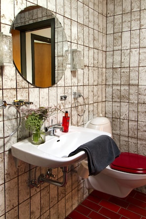 A bathroom with a white sink, a mirror, a toilet with a red seat, and a vase with flowers on the sink.