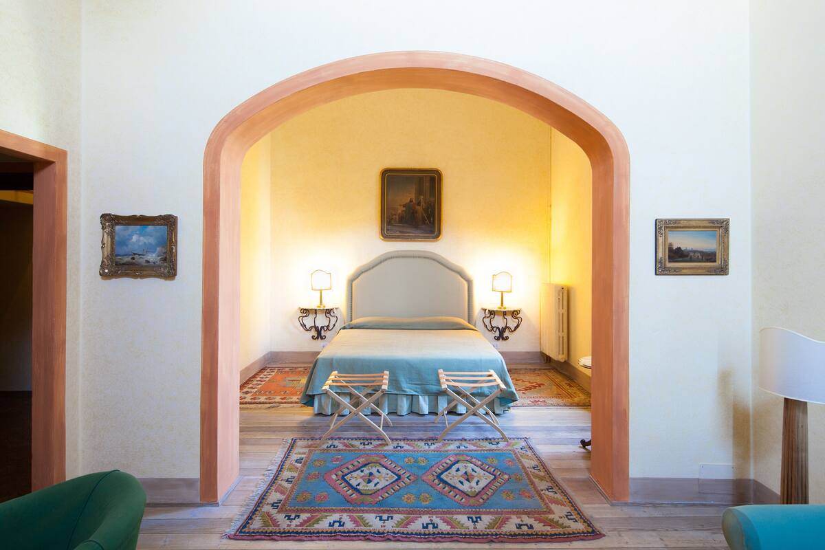 Un dormitorio con una cama, dos mesitas de noche, una lámpara auxiliar y una alfombra estampada, además de un arco de entrada.