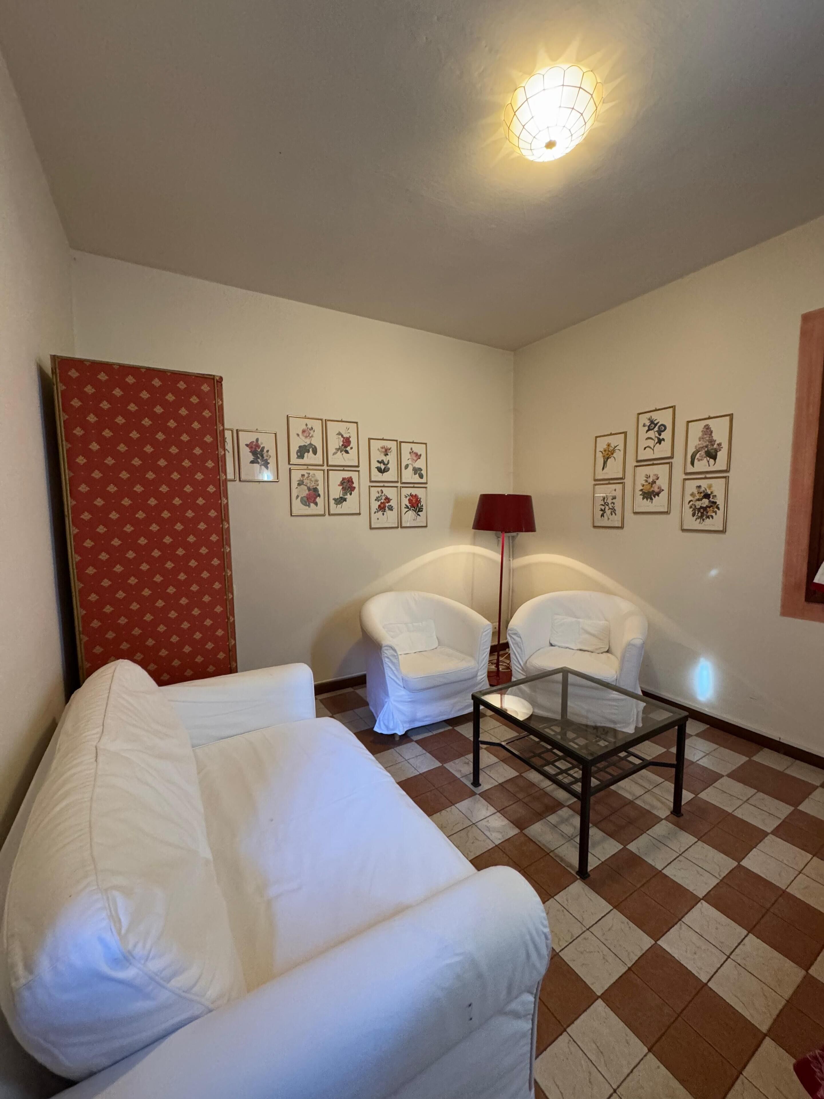 A living room with a checkered floor, white armchairs, a glass coffee table, and floral wall art.