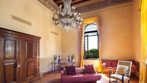 Una habitación amplia con un armario de madera, un sofá rosa con cojines estampados, un sillón blanco y un ventanal con cortinas amarillas.