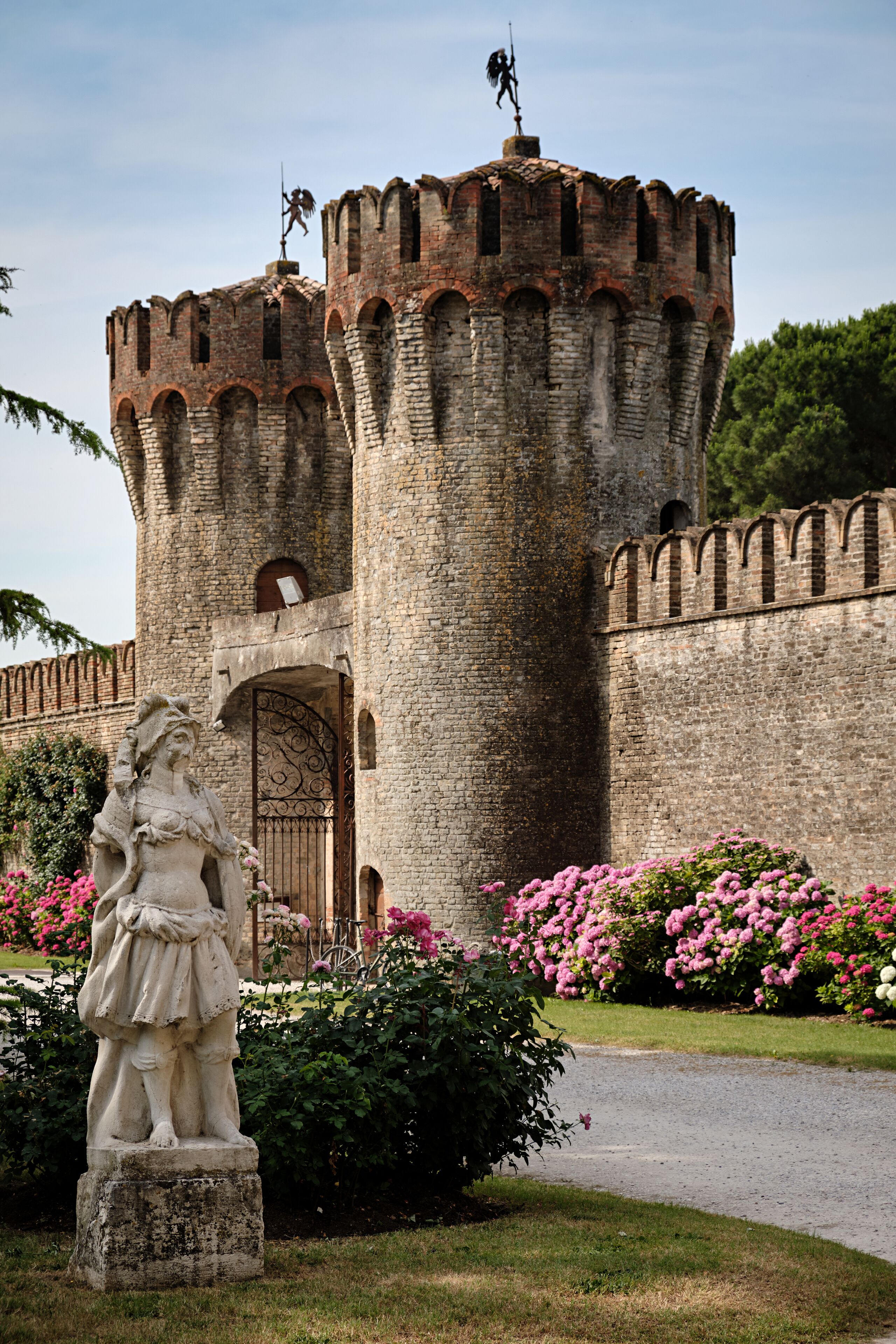 A historic stone castle with a central tower, a statue in the foreground, and a garden with pink flowers.