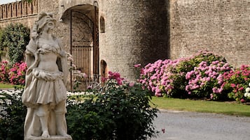 A historic stone castle with a central tower, a statue in the foreground, and a garden with pink flowers.