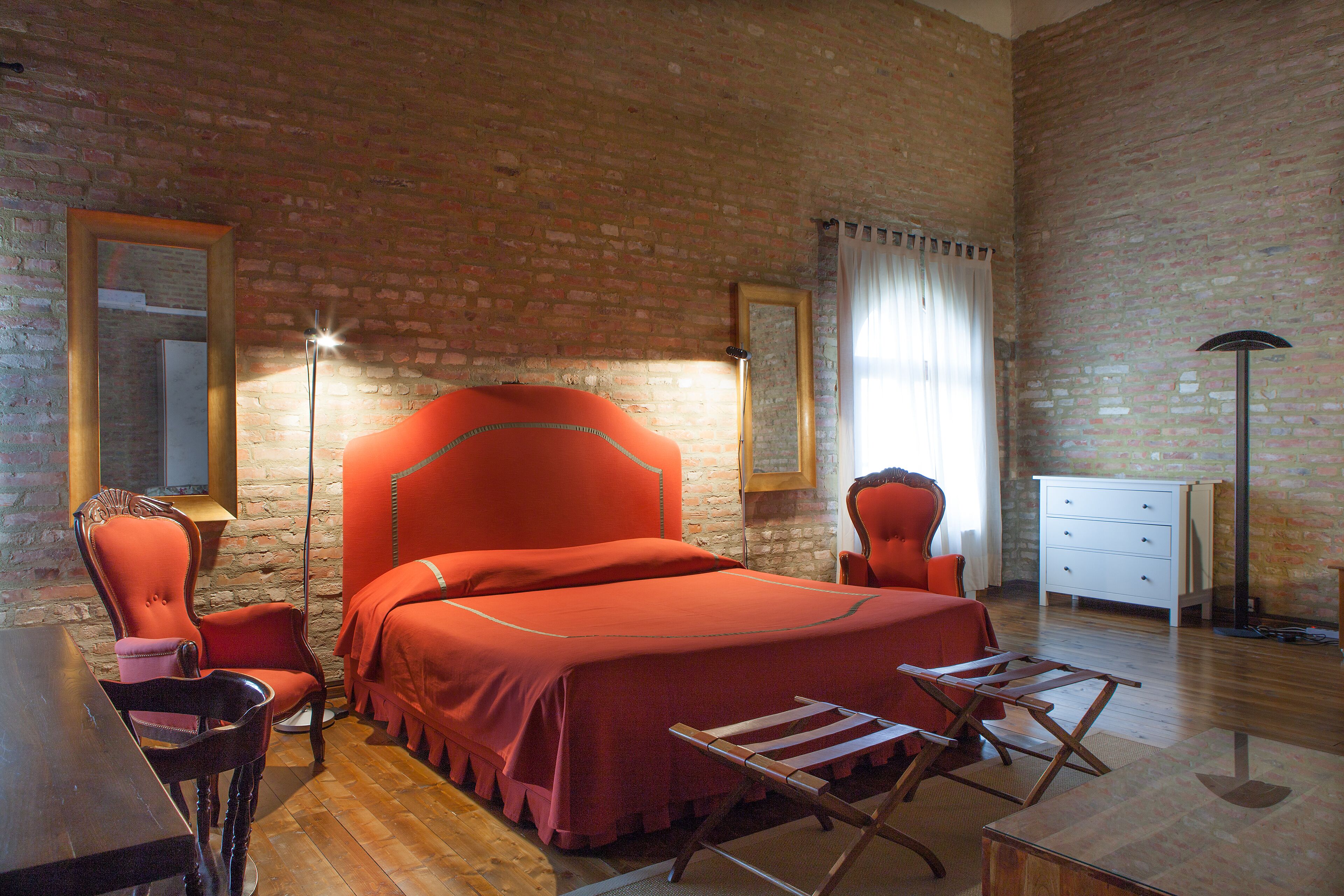 Een kamer met een bakstenen muur, een bed met een rode sprei, een houten bureau, een stoel, een kast en een vloerlamp.