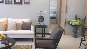 A living room with a white sofa, a black armchair, a small round table, and a coffee table. There are framed pictures on the wall and a potted plant on the side. - Baan Sarinya Pool Villa (Cha-am)