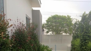 A well-maintained grassy area with a white building on the left, surrounded by various shrubs and a fence in the background. - Baan Sarinya Pool Villa (Cha-am)
