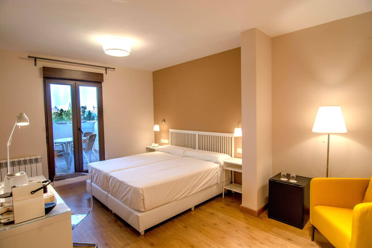 Una habitación de hotel con una cama, mesitas de noche, una silla, una lámpara y vistas a través de una puerta.