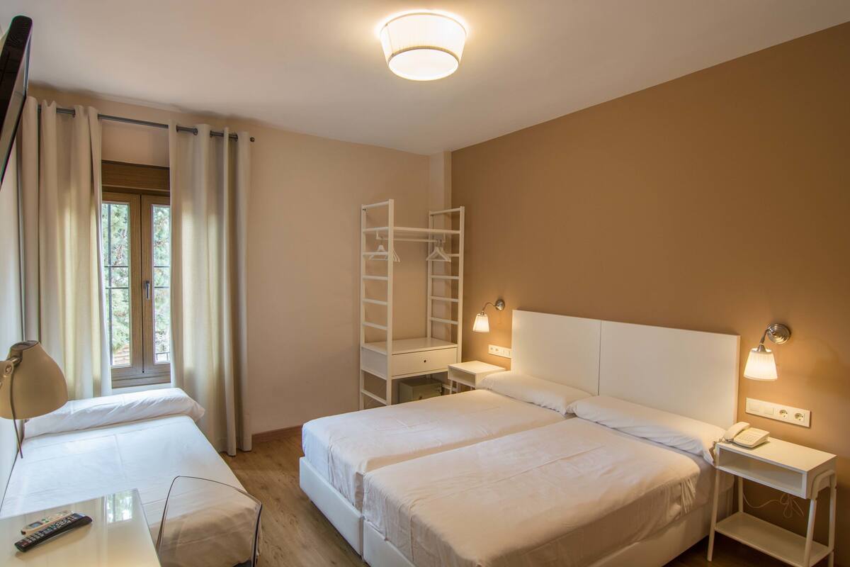Habitación de hotel con dos camas, un escritorio con lámpara, un armario y una ventana con cortinas.