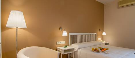 A hotel room with a bed, a chair, a bedside table with a lamp, and a tray with fruits and juice.